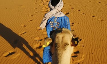 a man in a blue robe walking a camel in the desert