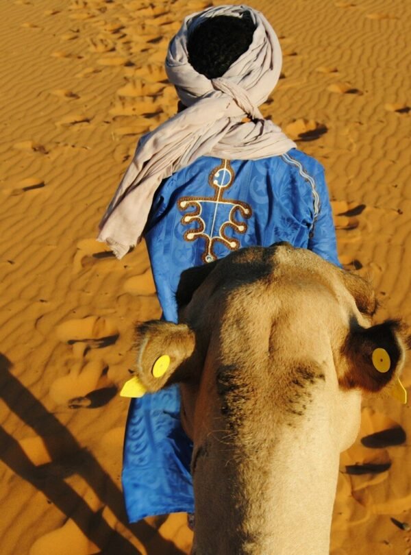 a man in a blue robe walking a camel in the desert